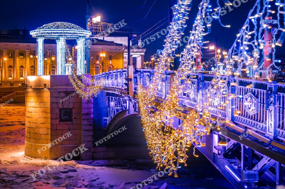Фотография рождественского и новогоднего центра Санкт-Петербург, Россия, сделанная зимой. На изображении показан Дворцовый мост, украшенный праздничными гирляндами и новогодней иллюминацией. Этот фотоснимок передаёт атмосферу Рождества и Нового года в Санкт-Петербурге: сияющие огни, зимний пейзаж и торжественный облик исторического центра города. Фото подчёркивает красоту мостов Санкт-Петербурга и праздничное оформление города в холодное время года. Иллюстрация может использоваться как изображение, картинка или слайд, посвящённые Рождеству, Новому году и зимним путешествиям по России.